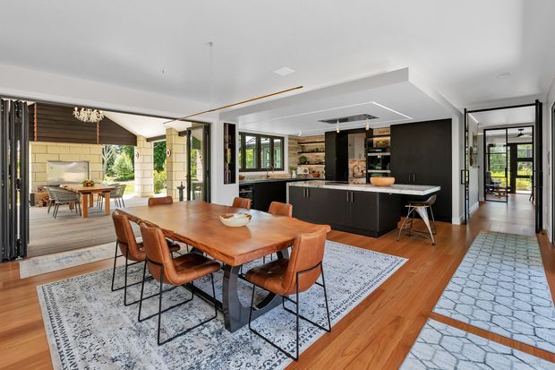 Open-plan contemporary kitchen, dining, and living space with warm timber flooring, a large solid timber dining table, black kitchen cabinetry and island, patterned rugs, and extensive black-framed glazing. The room opens toward a covered outdoor dining area and connects to a hallway beyond.