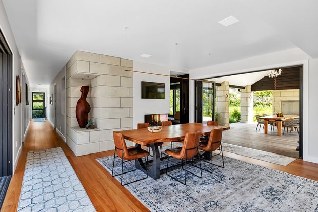 Bright open-plan dining area with a large live-edge timber dining table and leather dining chairs, positioned beside a substantial pale stone-clad feature wall with a recessed display niche and fireplace-like base. Warm timber flooring, patterned area rugs, dark-framed glazing, and a wide opening to a covered outdoor entertaining area are visible. The space has a contemporary, refined indoor-outdoor character.