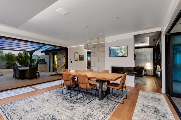 Open-plan contemporary dining and living area with a large timber dining table, leather dining chairs, patterned rugs, a fireplace, and wood flooring. The space opens directly to a covered outdoor entertaining area through expansive glazing and sliding doors.