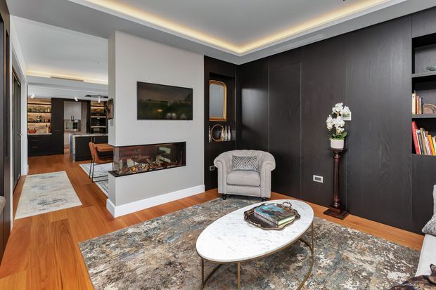 A refined contemporary living room with dark timber wall panelling and built-in shelving, a white feature fireplace wall with inset firebox and television, an upholstered armchair, oval stone coffee table, and a large patterned area rug. Warm timber flooring and concealed perimeter lighting create a sophisticated, layered interior. A kitchen and dining area are visible through the opening at left.