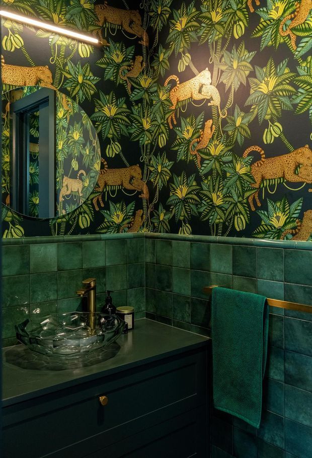 A richly decorated compact bathroom or powder room with deep green square wall tiles, a dark green vanity with a transparent vessel sink and brass tap, and a large mirror. The upper walls feature dramatic dark botanical wallpaper with repeated golden leopards and lush green foliage. A teal towel hangs from a brass rail, creating a dark, theatrical and luxurious atmosphere.