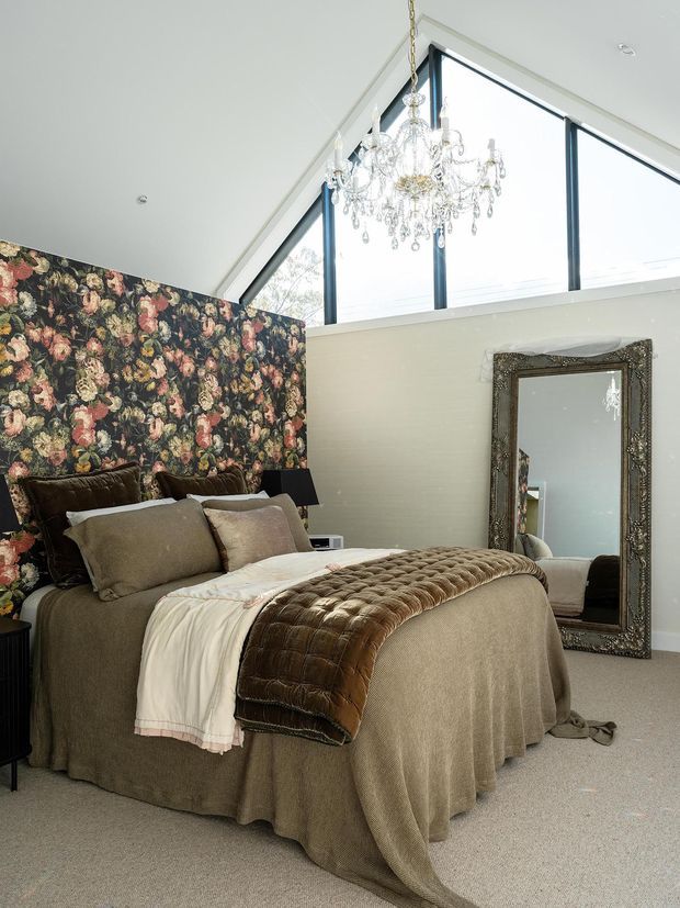 A sophisticated bedroom with a layered taupe and cream bed, plush pillows, a quilted brown throw, and a dark floral feature wall. A large ornate floor mirror leans against the opposite wall, while a crystal chandelier hangs beneath a dramatic high triangular window. The room has soft neutral finishes, carpeted flooring, and abundant natural light.