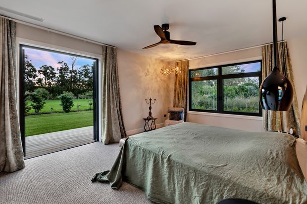 A warm, refined bedroom with a large bed dressed in a muted sage-green cover, soft carpeted flooring, textured plaster walls, patterned curtains, and black-framed windows. A sliding glass door opens toward a lush green garden, while a sculptural pendant light and ceiling fan add visual interest.