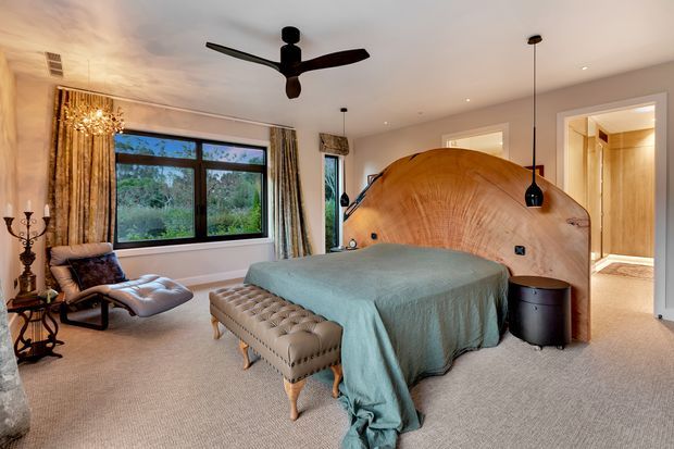 A spacious, warmly lit bedroom with a large upholstered bed framed by a sculptural curved timber headboard. The room features wall-to-wall carpeting, a tufted bench, a leather lounge chair and ottoman, dark pendant lights, patterned curtains, and a black ceiling fan. A bathroom or dressing area is visible through the open doorway.