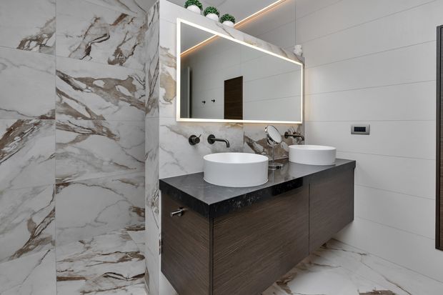 Contemporary bathroom with a floating dark timber vanity, two white vessel basins, a black stone benchtop, and a large illuminated rectangular mirror. Dramatic white marble-look wall and floor tiles with warm veining contrast with pale grey wall panels and matte black tapware and hardware.