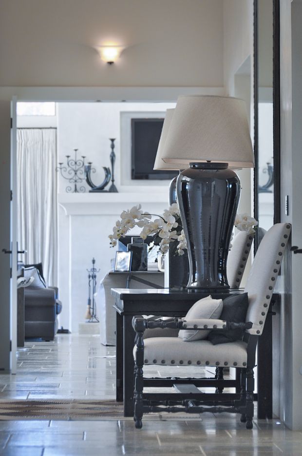 A refined interior view looking along a polished tiled hallway or transitional space. In the foreground is a dark wood console table with a large glossy black ceramic lamp, cream shade, framed photographs, and white orchid arrangement. A button-tufted cream upholstered armchair sits beside it. The background shows a bright sitting area with a fireplace mantel, decorative accessories, and a dark wall-mounted screen. The space has white walls, dark trim, and a restrained, elegant monochromatic palette.