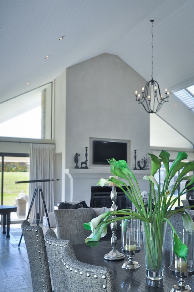 Bright, elegant open-plan living and dining space with a high vaulted white paneled ceiling, large windows, and a central fireplace with a pale mantel. A dark dining table is surrounded by upholstered nailhead-trimmed chairs and decorated with tall green botanical arrangements. A black chandelier hangs above, while a lounge area and outdoor landscape are visible beyond the glazing.
