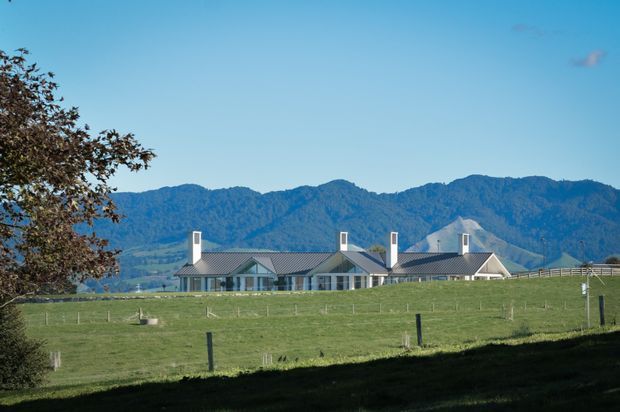 A long, low rural property set behind expansive fenced pasture, with multiple connected gabled roof forms, pale exterior walls, dark grey metal roofing, and several prominent chimneys. Forested mountains rise behind the building under a clear blue sky, creating a distinctly alpine or rural landscape setting.