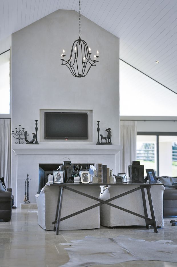 A refined open-plan living and dining space centred on a tall white plaster fireplace wall with a mounted television and an ornate black chandelier. A narrow console table with crossed metal supports separates the dining area from the seating area, with upholstered dining chairs, sofas, framed photographs and decorative accessories. The room features a high pitched white paneled ceiling, large windows, pale stone flooring and a light neutral palette.