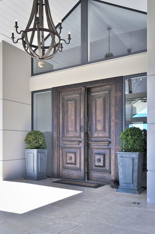 A grand covered entryway with tall, dark timber double doors featuring ornate raised panel detailing. Pale rendered walls and a white-lined vaulted ceiling surround the entrance, with dark-framed clerestory glazing above and beside the doors. A large wrought-iron candle-style chandelier hangs overhead, while two clipped spherical topiary plants sit in tall grey square planters. The floor is finished in large grey stone-like tiles, creating a formal, refined entrance.