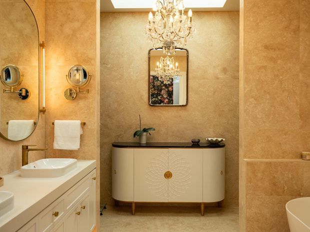 Elegant luxury bathroom featuring warm beige stone-effect wall and floor finishes, a freestanding vanity with cream cabinetry and dark stone benchtop, twin wall-mounted basins and brass fixtures, a large mirror, glass shower enclosure, chandelier, and a partially visible freestanding bathtub.