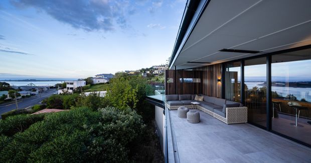 Contemporary multi-level coastal residence viewed from an elevated outdoor terrace, with expansive glazing, dark timber-clad walls, a broad overhanging roof, and an outdoor lounge overlooking landscaped gardens, neighbouring homes, and a distant body of water. The terrace uses large pale stone or concrete-look paving and includes built-in seating, a coffee table, and woven ottomans.
