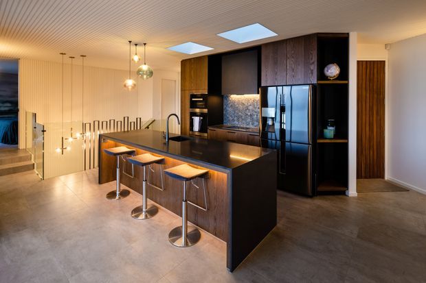 A contemporary open-plan kitchen dominated by a large dark island with seating for four. The kitchen features black and dark timber cabinetry, a reflective dark refrigerator, integrated appliances, a patterned backsplash, and warm under-cabinet lighting. Pendant lights hang above the island, while linear grooved ceiling and wall detailing, skylights, timber screening, and a stair connection add architectural interest. The space has polished concrete-look flooring and a warm, refined atmosphere.