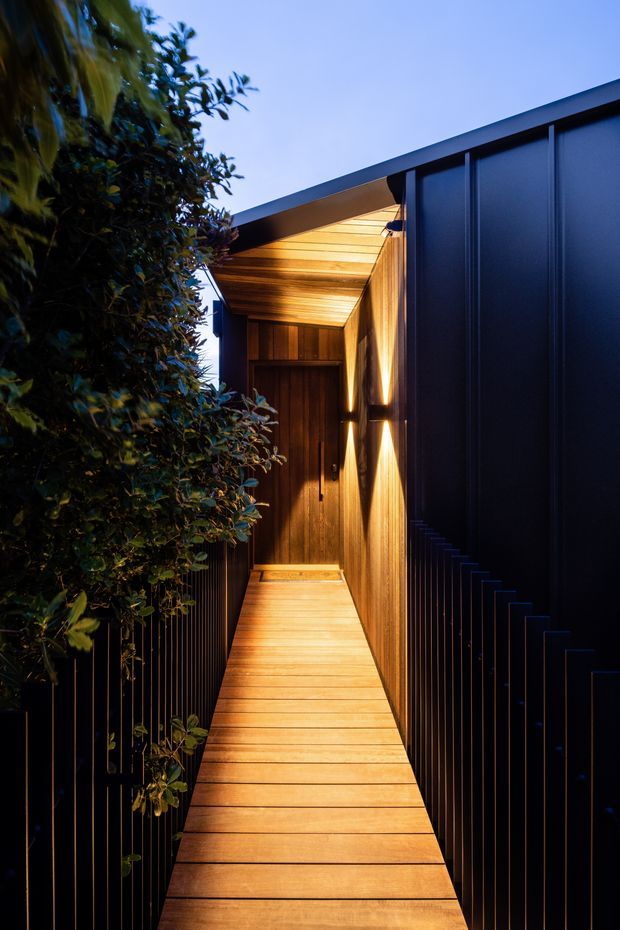 A narrow, sheltered exterior walkway leading toward a timber-clad entry door. Warm linear lighting illuminates the timber decking, soffit, and vertical timber wall, while dark metal-clad walls and slender black railings frame the passage. Dense green planting borders the left side, creating a dramatic, intimate approach at dusk.