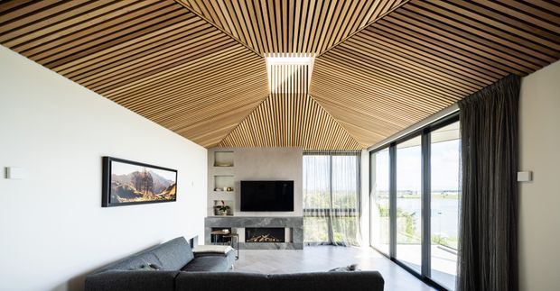 Contemporary living room with a dramatic geometric timber batten ceiling, white walls, dark-framed floor-to-ceiling glazing, a fireplace and television wall, and a dark sectional sofa. The glazing opens toward a bright waterfront outlook, while sheer and dark curtains provide contrast.