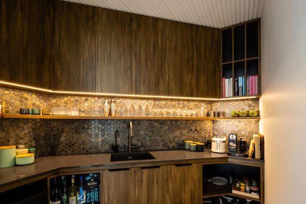 A compact, warmly lit contemporary kitchen with dark timber upper and lower cabinetry, a dark stone-look benchtop, and a distinctive hexagonal mosaic tile splashback. Integrated warm LED strip lighting illuminates open shelving displaying glassware and books. The kitchen includes a sink, cooktop, small appliances, open storage, and a ribbed white ceiling, creating a rich, moody and refined atmosphere.