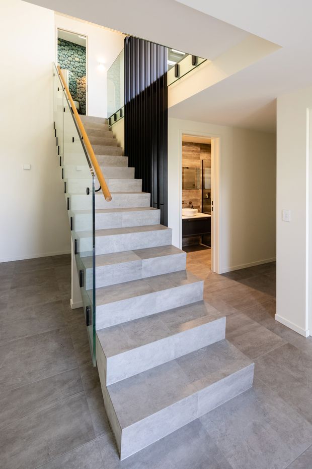 Contemporary interior stairway with broad grey tiled steps, clear glass balustrades, a timber handrail, and a dramatic black vertical-slat screen. The stair rises through a bright open landing, with a partially visible bathroom doorway and warm timber finishes beyond.