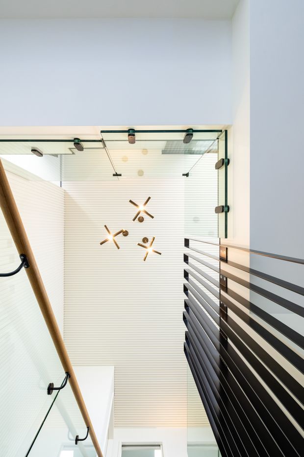 Upward-looking view of a contemporary interior stairway or landing. A black horizontal metal balustrade runs along the right side beside a glass balustrade with timber handrail on the left. The space has white walls, a white linear-panel ceiling, exposed glass detailing, and several modern sculptural pendant lights.