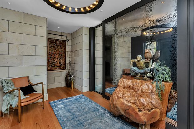 A refined interior hallway or entry space with pale large-format wall tiles, warm timber flooring, a blue area rug, a mid-century leather lounge chair, decorative artwork, and a dramatic black-framed textured mirrored glass partition. A sculptural timber console with seated figures and foliage forms a focal point, while a circular statement light is reflected in the glazing.