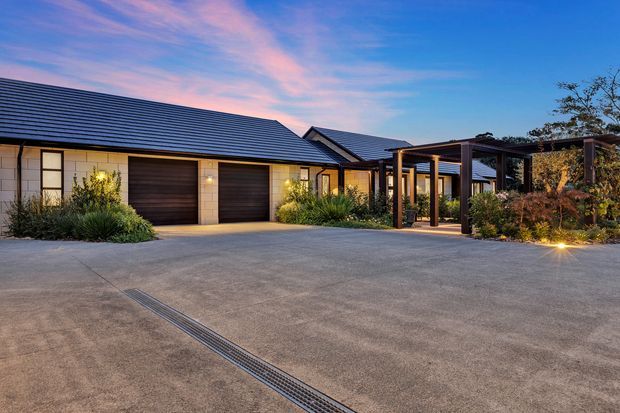 A single-storey contemporary residential property viewed from a broad concrete driveway at dusk. The building has dark pitched tiled roofs, pale masonry walls, two dark timber garage doors, and a covered timber-framed entry or outdoor walkway with landscaped planting and warm exterior lighting.