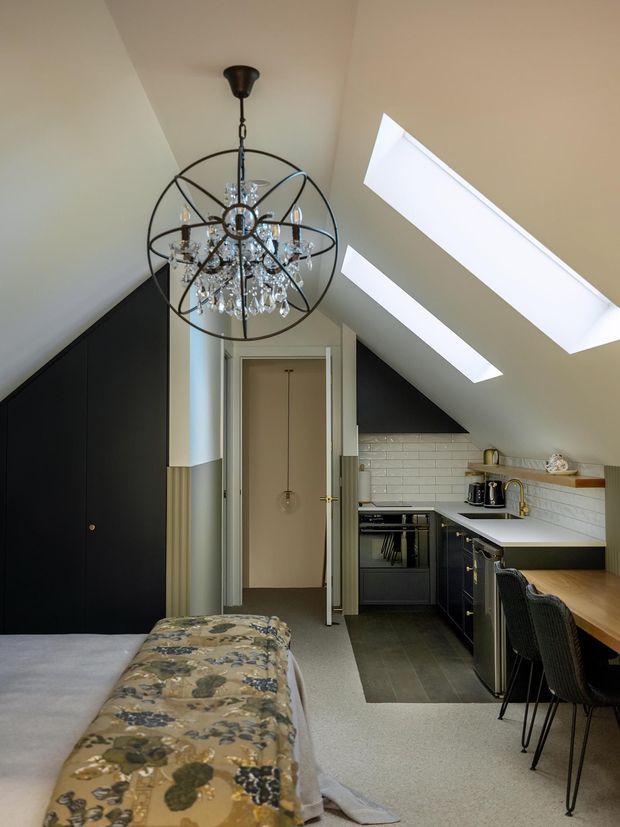 A compact open-plan attic interior combining a bedroom in the foreground with a small kitchen and dining/work counter at the rear. The space has a steep sloping ceiling with two large skylights, dark built-in cabinetry along the left wall, a white subway-tile splashback, pale benchtops, dark flooring in the kitchen zone, and a dramatic spherical crystal chandelier.