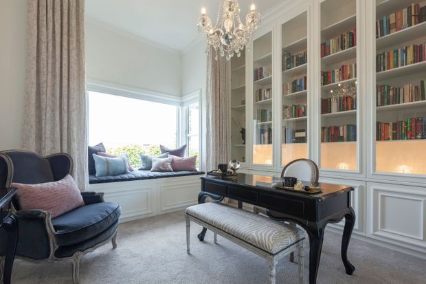 Elegant traditional-style reading room or library with floor-to-ceiling built-in bookcases, a black lacquered console desk, upholstered bench, ornate armchairs, a window seat, patterned curtains, and a crystal chandelier. The room has pale wall panelling, soft carpeting, and abundant natural light.