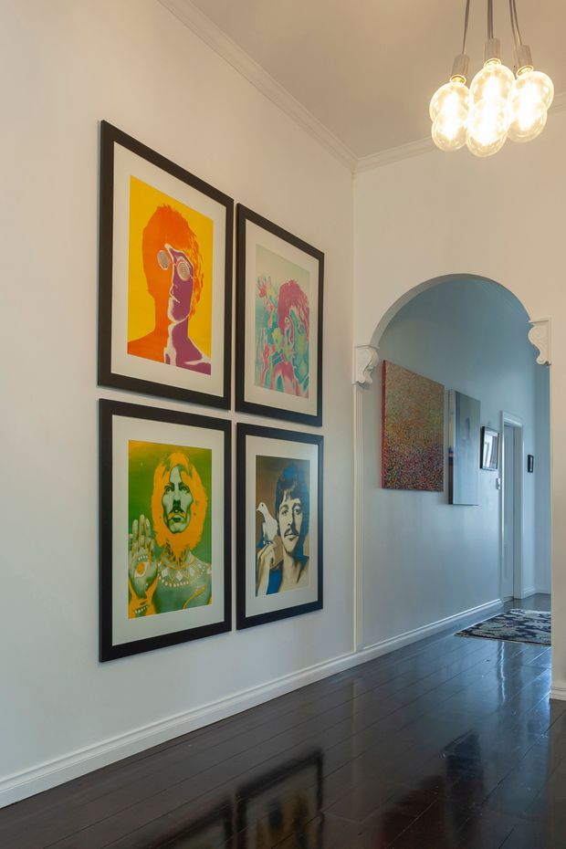 A refined residential hallway with dark polished timber flooring, pale walls, decorative cornices and skirting, and a softly arched opening with ornamental trim leading to another corridor. Four colourful framed artworks form a gallery wall, while a cluster of glowing pendant lights hangs overhead. The space combines classic architectural detailing with bold contemporary art.