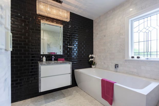 A stylish contemporary bathroom featuring a freestanding white bathtub, wall-mounted white vanity, black glossy subway-tile feature wall, pale textured wall tiles, a decorative leaded-glass window, and a rectangular pendant light. Small mosaic floor tiles and pink towels add contrast.