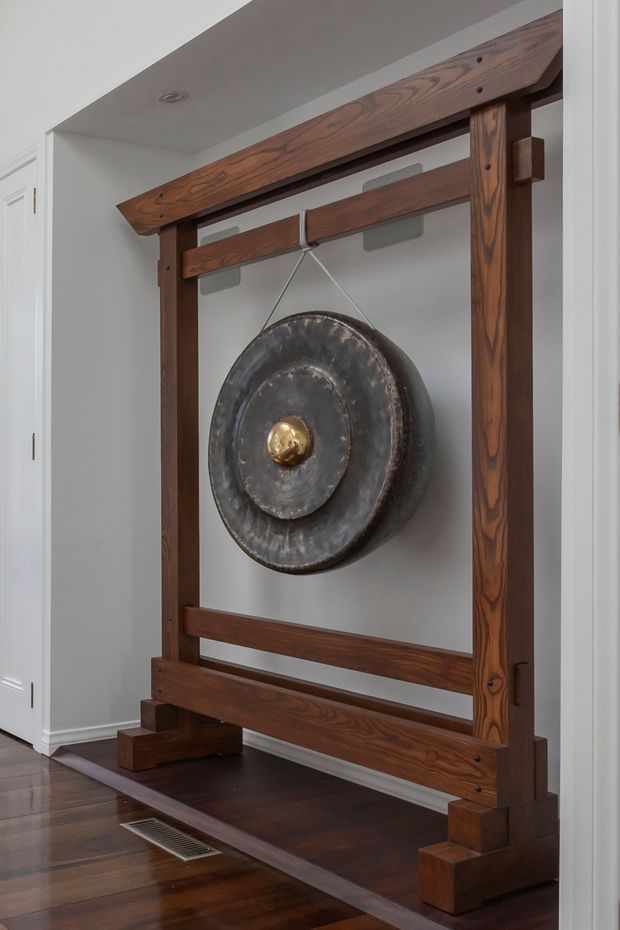 Interior view of a large suspended metal gong mounted within a substantial dark timber frame. The custom frame has exposed joinery, horizontal rails, and stepped timber feet, set against pale painted walls with white trim and dark timber flooring in a hallway or entry area.