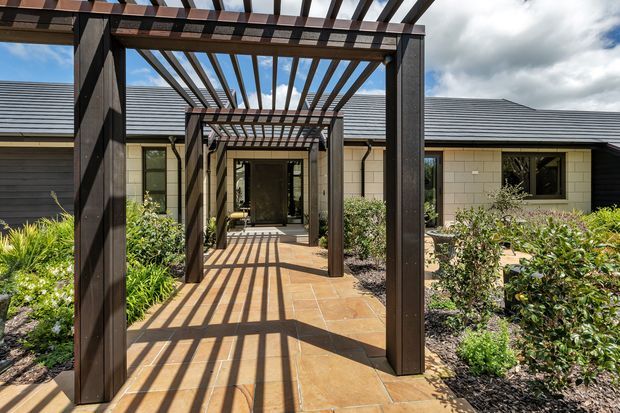 Exterior view of a low-rise contemporary home entry, approached along a paved walkway framed by a series of dark timber pergolas. The pergola slats cast strong linear shadows across the warm-toned paving, with landscaped garden beds on either side. The house features dark horizontal cladding, pale masonry walls, charcoal hipped roofing, and large dark-framed windows.