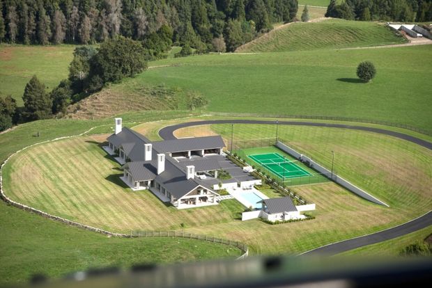 Aerial view of a large estate-style property set within expansive rolling green farmland. The complex includes several connected pitched-roof buildings with dark roofing and light exterior walls, extensive manicured lawns, curved paved driveways or paths, and a fenced green tennis court. Mature trees and open rural fields surround the site.