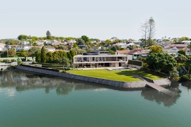 Aerial exterior view of a contemporary multi-level waterfront residence on a landscaped peninsula, with extensive glazing, horizontal rooflines, stone retaining walls, a broad lawn, and direct water frontage. A suburban neighbourhood and mature trees extend behind the property.