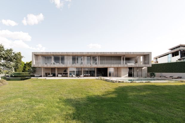 A wide exterior view of a contemporary two-storey residence set behind an expansive lawn. The building features a long rectangular form, strong horizontal lines, extensive floor-to-ceiling glazing, deep roof overhangs, slender vertical screens, and a covered ground-level terrace. A swimming pool or narrow reflecting pool appears along the front, with neighbouring modern housing visible at the right.