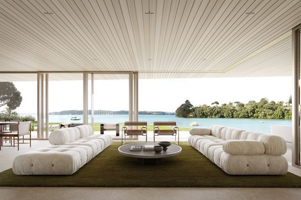 A bright, expansive waterfront living room with floor-to-ceiling glazing opening directly to a tranquil bay. Two large, low-profile cream modular sofas face each other across a dark olive-green rug and round coffee table. A ribbed white ceiling with recessed spotlights, slender timber-toned window frames, and a minimal arrangement of mid-century-inspired furniture create a refined, relaxed interior overlooking the water.