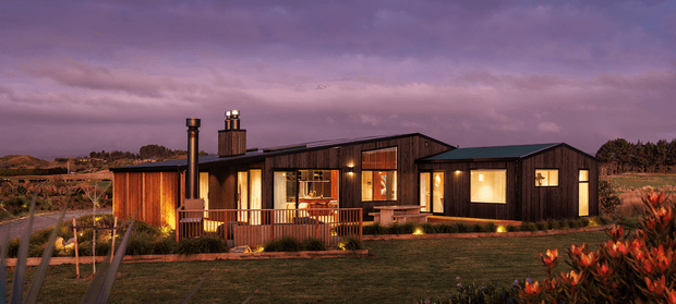 A single-storey contemporary rural home photographed at dusk, with dark timber cladding, warm illuminated windows, a low pitched roofline, covered timber veranda, and landscaped lawn. The house sits in an open rural setting with planting around the foreground and subdued purple evening sky.