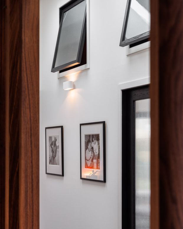 A tall, narrow interior hallway or entry passage viewed through dark timber door frames. White walls rise to a high ceiling and feature black-framed textured-glass windows and a matching black-framed glazed door. A modern wall sconce illuminates two framed artworks, creating a refined monochrome palette with warm wood accents.