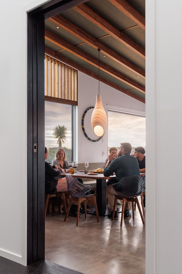 A contemporary dining area viewed through a wide dark-framed doorway. The room features a long dining table with upholstered and transparent chairs, a sculptural pendant light, large windows, polished concrete flooring, and a dramatic ceiling with exposed timber beams and dark acoustic panels. Several people are seated at the table, with a palm tree and coastal landscape visible outside.