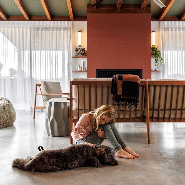 Warm, timber-framed living room with a dramatic exposed-beam ceiling, polished concrete floor, full-height sheer curtains, and a central red brick fireplace. A mid-century-style timber sofa faces the fireplace, with built-in shelving and soft neutral furnishings. A child and dog are present in the foreground.