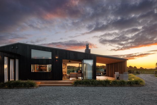 Single-storey contemporary home at sunset, clad in dark vertical board-and-batten-style siding with extensive glazed openings. The composition includes a projecting covered outdoor living area, timber soffit, warm interior lighting, a chimney, timber entry deck, gravel forecourt, and surrounding low landscape. The flat or very low-pitched roof and horizontal pavilion-like form are clearly visible.