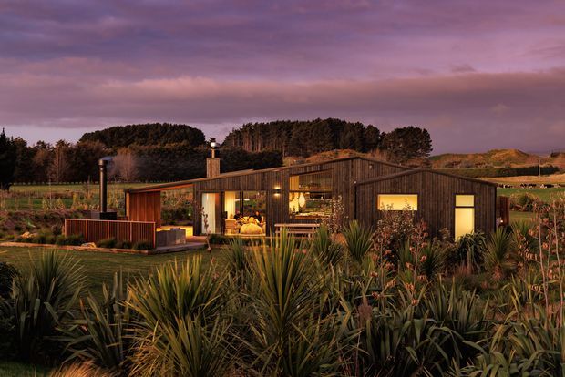 A single-storey contemporary rural dwelling set within a dense landscaped garden. The building has dark timber-clad walls, a low pitched roof, extensive glazing, warm interior lighting, and a small sheltered outdoor deck or terrace. The surrounding planting and rolling rural landscape are shown at dusk beneath a dramatic purple sky.