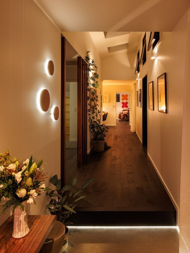 A warmly lit residential hallway viewed from an entry area, with dark timber flooring, cream painted walls, recessed ceiling details, framed artwork, decorative wall sconces, a large floral arrangement, potted plants, and a softly illuminated step leading into the corridor. The hallway extends toward a brighter room at the far end.
