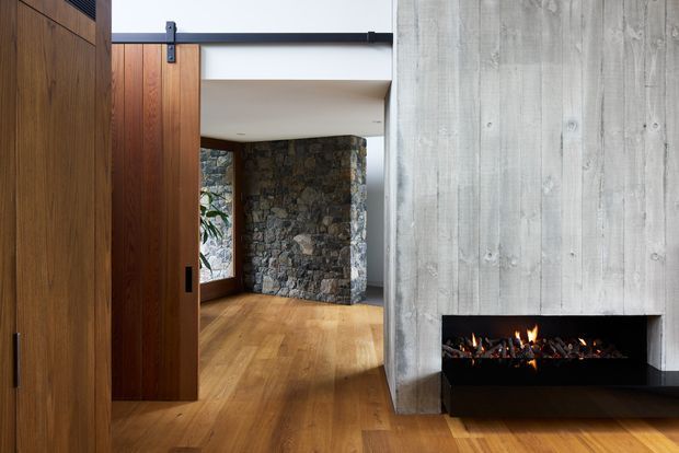 Interior view of a modern entry or circulation space with a large timber pivot door and exposed black sliding hardware. Warm timber flooring and vertical timber wall panelling frame a rough natural-stone feature wall, while a concrete-look fireplace wall with a long built-in fire creates a strong focal point. The space has a restrained, material-focused contemporary character.