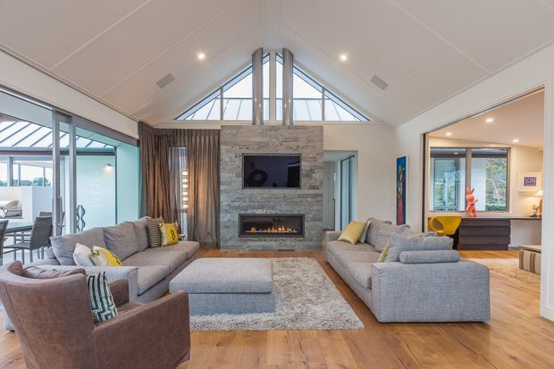 A bright, spacious contemporary living room with a vaulted ceiling and triangular clerestory glazing. Two large grey sectional sofas and upholstered armchairs surround a central ottoman and rug, facing a stone-clad feature wall with a television and built-in fireplace. Full-height glazing and open connections lead to adjacent dining and kitchen areas. Warm timber flooring contrasts with the cool grey upholstery and pale walls.