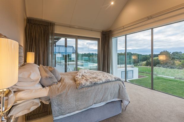 A warm, contemporary bedroom with a bed positioned beside expansive floor-to-ceiling glazed sliding doors. Neutral bedding, textured cushions, full-length taupe curtains, and carpet create a soft, calm interior. The glazing opens onto a landscaped lawn and pool area, with a view toward a modern glazed structure outside. Recessed ceiling lighting and bedside lamps provide gentle illumination.