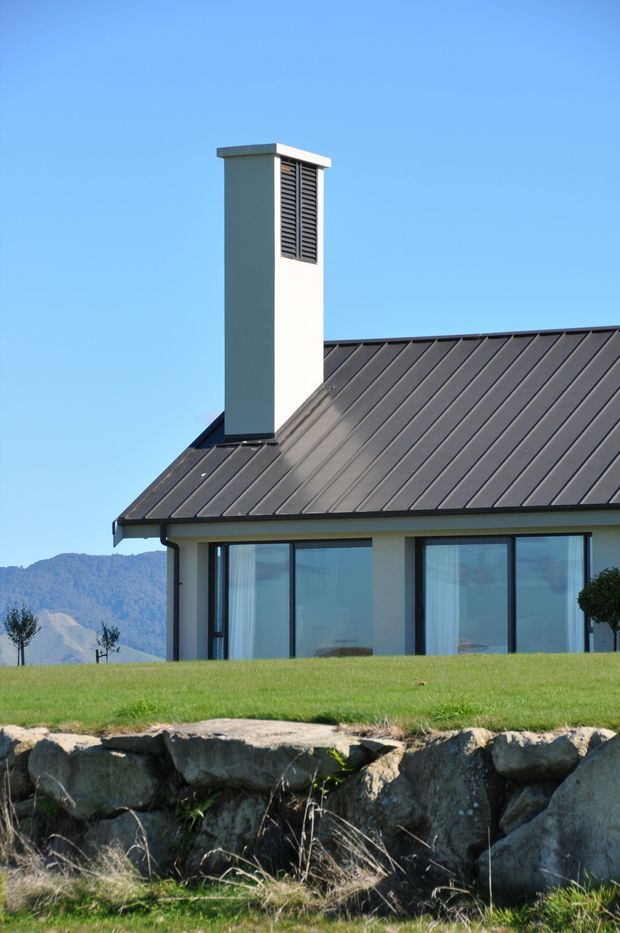 Exterior close-up of a contemporary single-storey house, focused on a dark standing-seam pitched roof, white rendered chimney with a louvred vent, and large dark-framed windows. The house sits above a rough stone retaining edge with a mountain landscape visible in the background under a clear blue sky.