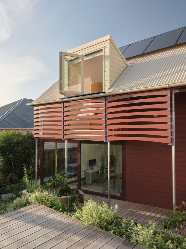 Exterior view of a contemporary multi-level home with a steep corrugated metal roof, dark solar panels, red-brown weatherboard cladding, and a distinctive upper-level dormer window. A curved horizontal timber privacy screen projects over glazed ground-floor living spaces, opening onto a timber deck and landscaped garden.