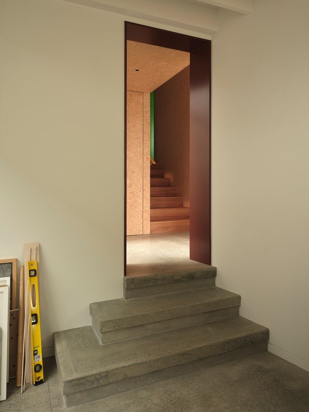 Interior view of a compact stairway leading through a dark timber-framed doorway into a warm timber-lined stairwell. The foreground steps are raw grey concrete, while the inner stair treads and walls are timber-toned, with a muted pale wall surrounding the opening.