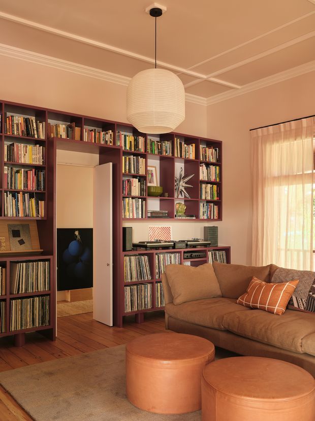 Warm, richly coloured living room featuring extensive dark burgundy built-in bookshelves filled with books and vinyl records, a turntable and stereo equipment, a large brown sectional sofa, leather ottomans, timber flooring, and a central oversized paper pendant light. Decorative ceiling mouldings and coffered detailing frame the room, while sheer curtains filter daylight through a tall window or glazed door.