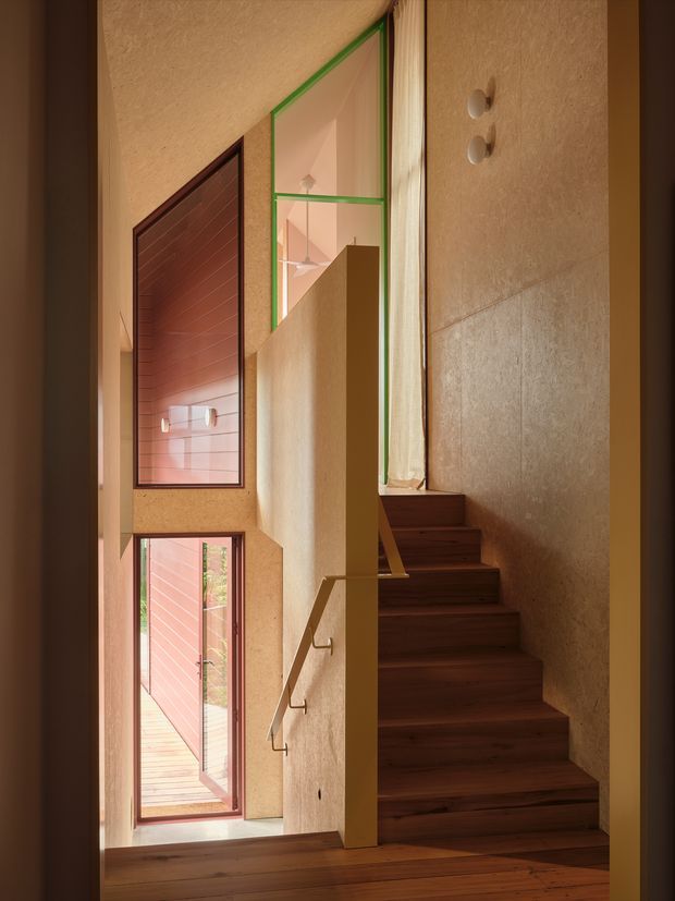 A tall, warm-toned interior stairway with dark timber treads and landing, a simple timber handrail, textured tan plaster walls, and a green-framed clerestory window. A glazed opening and red-toned exterior door are visible along the stair hall, emphasizing the vertical circulation space and natural light.