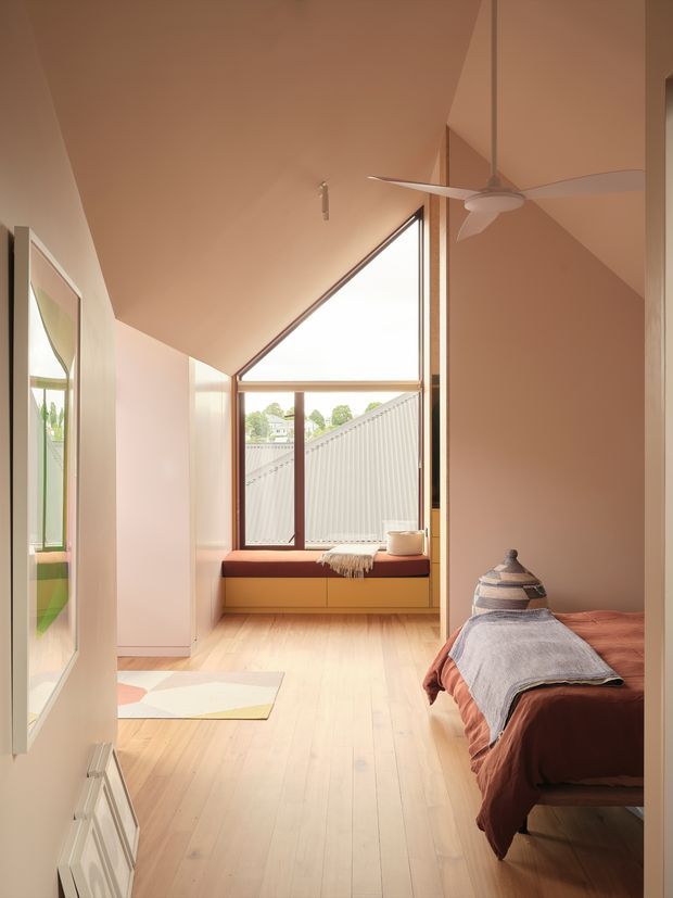 Warm, softly styled bedroom with a sloping ceiling and dramatic triangular window overlooking a neighbouring roofline. The room features pale timber flooring, peach-toned walls and ceiling, a built-in window seat with drawers, a ceiling fan, and a bed with muted rust and grey bedding. A large framed mirror reflects greenery and part of the room.